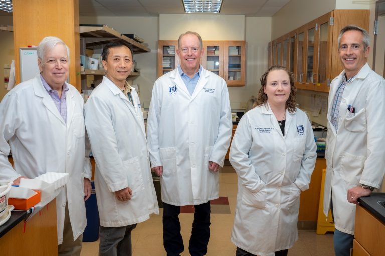 Five people in white coats stand in middle of lab