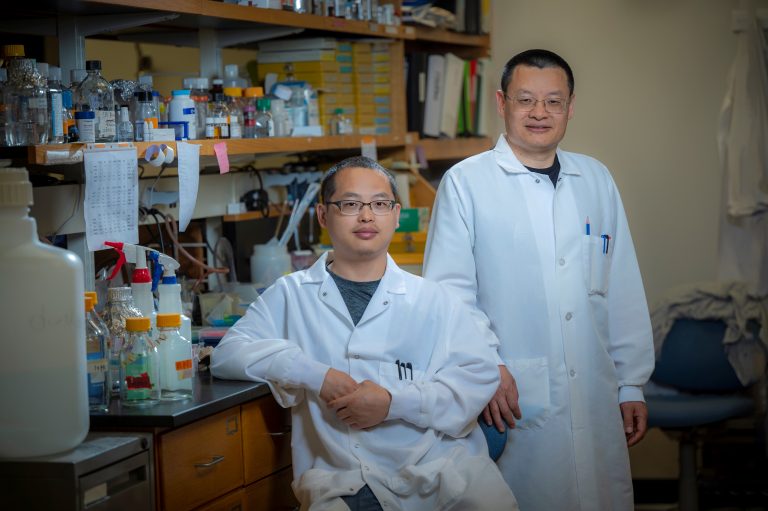 Two men in white coats sit in front of lab