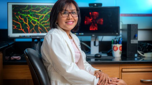 Woman in white coat sits in front of computer screens with cellular images on them