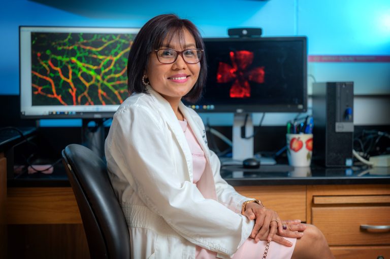 Woman in white coat sits in front of computer screens with cellular images on them