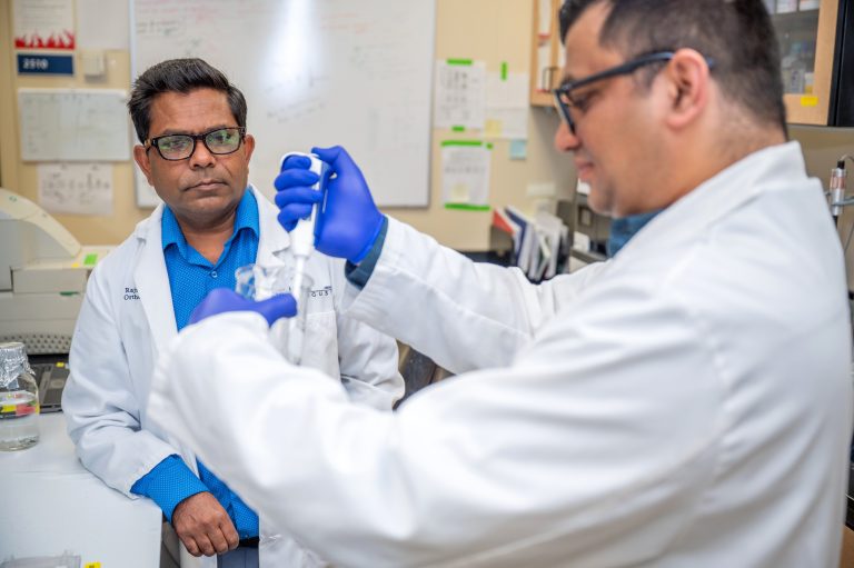 Man in white coat and blue shirt watches and man with gloves pipes liquid into tube