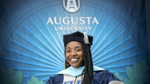 Dr. Shareen Clement wearing graduation regalia while standing in front of Augusta University sign on the Summerville Campus.