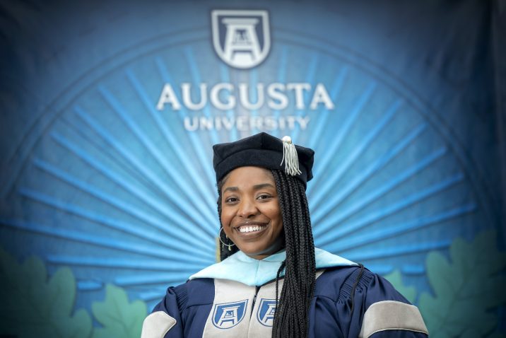 Dr. Shareen Clement wearing graduation regalia while standing in front of Augusta University sign on the Summerville Campus.