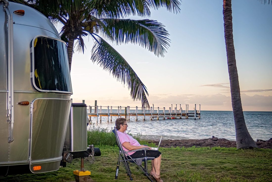 woman sits outside her Airstream with a computer on her lap doing work near the ocean
