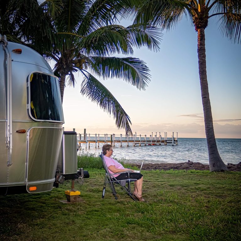 woman sits outside her Airstream with a computer on her lap doing work near the ocean