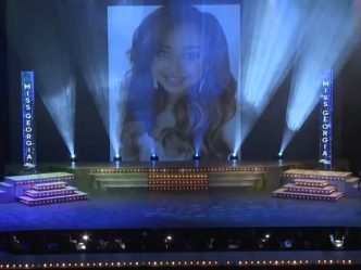 a woman's face appears on a large screen over a pageant stage