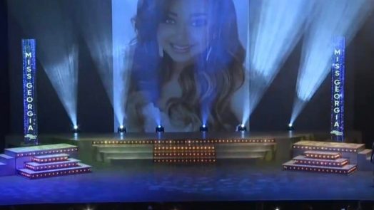 a woman's face appears on a large screen over a pageant stage