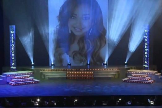 a woman's face appears on a large screen over a pageant stage