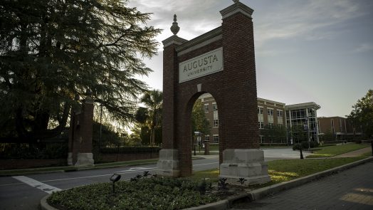 The entrance sign to Augusta University on the Summerville campus with the sun setting to the left of it behind a tree