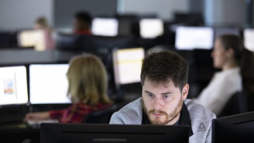 A man sits at his computer. Behind him, various people sit at other computers in a computer lab.