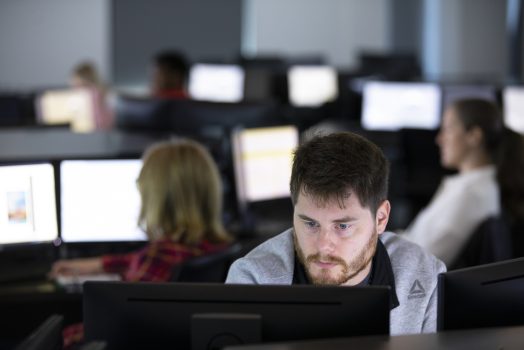 A man sits at his computer. Behind him, various people sit at other computers in a computer lab.