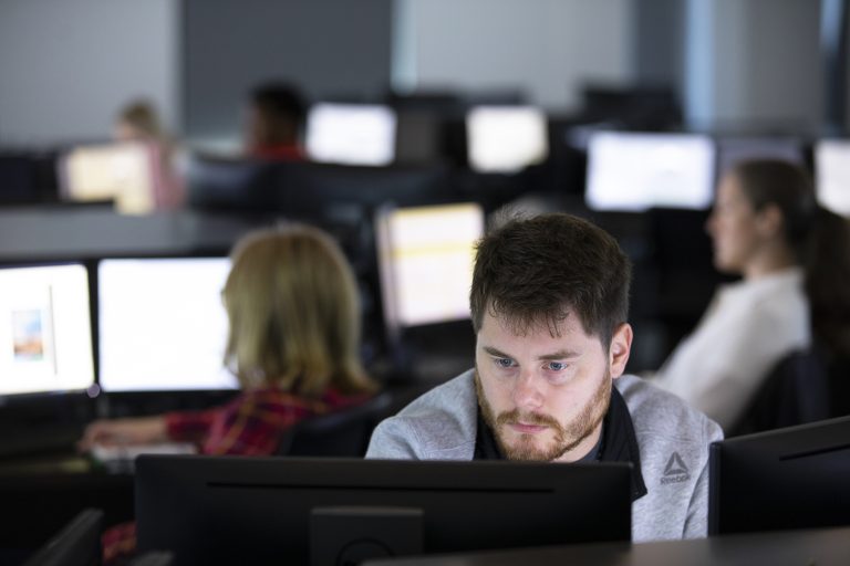 A man sits at his computer. Behind him, various people sit at other computers in a computer lab.