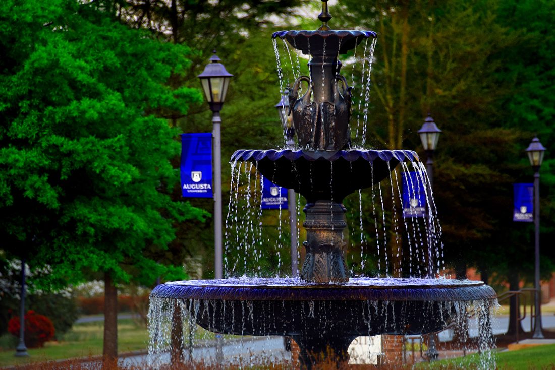 water cascades down from a three-tiered fountain on AU campus with blue AU flags on flagpoles and trees cover the background