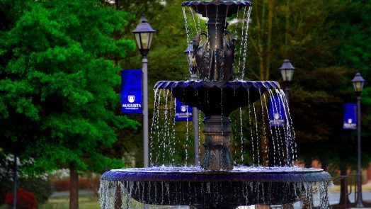 water cascades down from a three-tiered fountain on AU campus with blue AU flags on flagpoles and trees cover the background