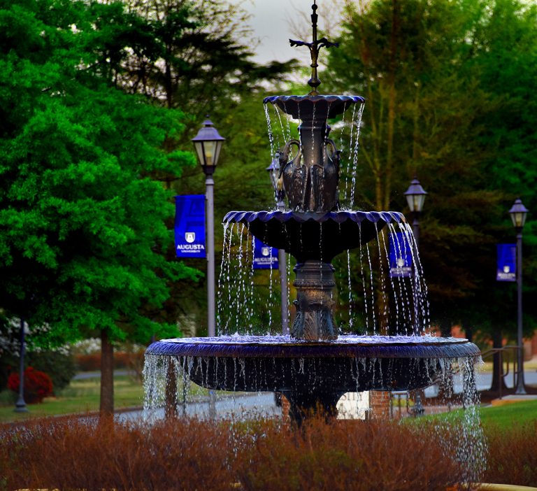 water cascades down from a three-tiered fountain on AU campus with blue AU flags on flagpoles and trees cover the background