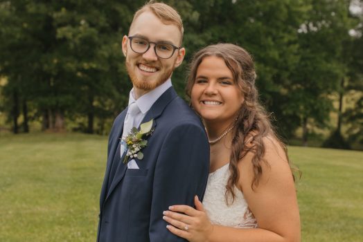 a bride stands behind her husband as they smile at the camera on their wedding day