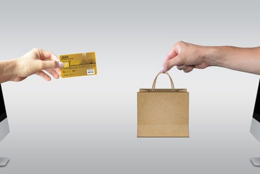 a computer with a hand holding a credit card coming out of it faces a computer with a hand holding a shopping bag coming out of it