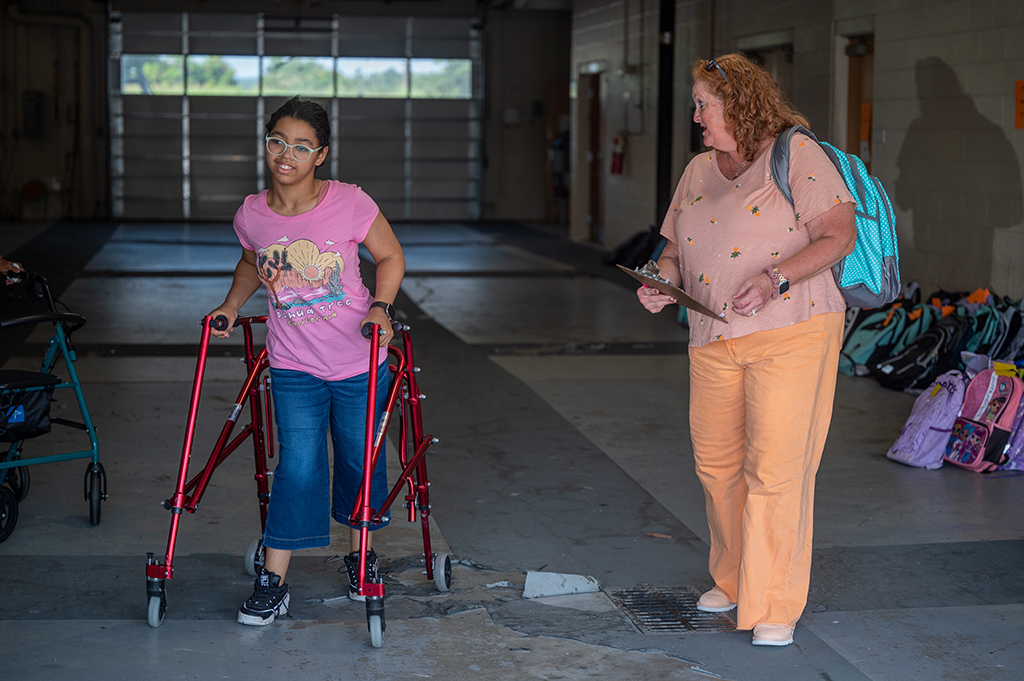 Girl using a walker with a woman holding her backpack.