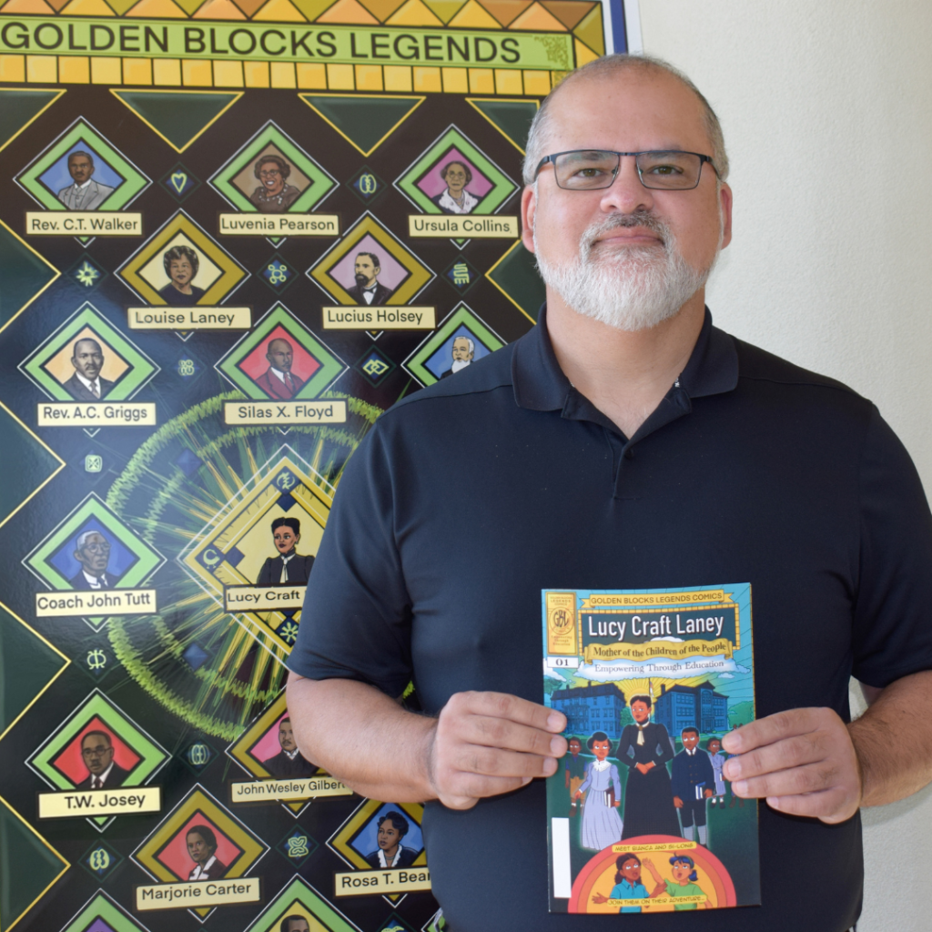 Dr. Juan Walker wearing a black shirting standing in front of Golden Blocks Legend Poster and holding Golden Blocks Legends Comic book.