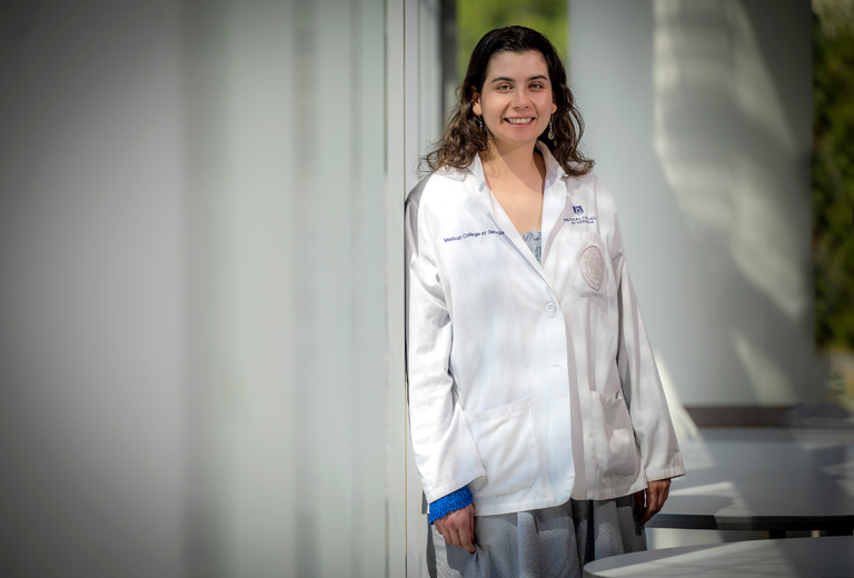 Woman wearing doctor's lab coat stands against a column while smiling for a picture.