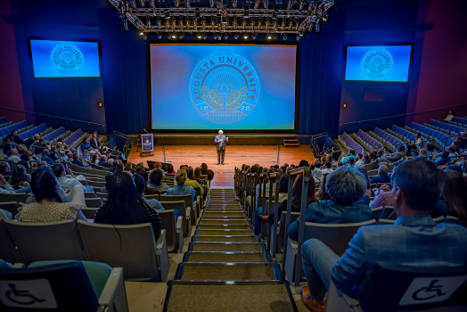 A man in a suit presents to a large group in front of a projection screen in a theater.