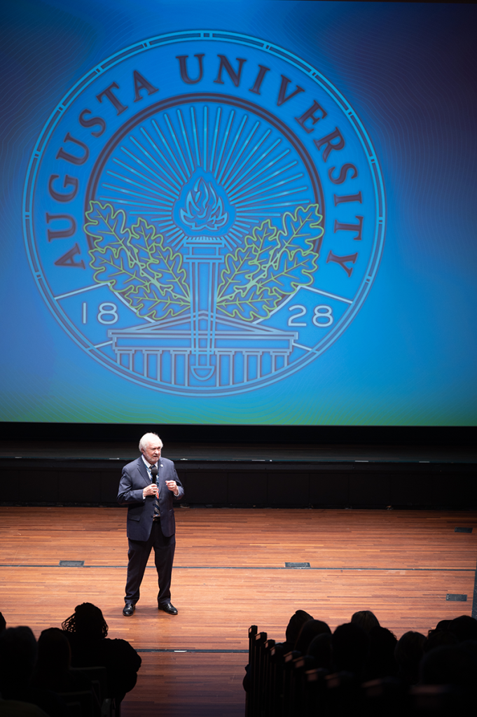 A man in a suit talks into a microphone while standing on a stage in front of a large image of a seal for Augusta University.