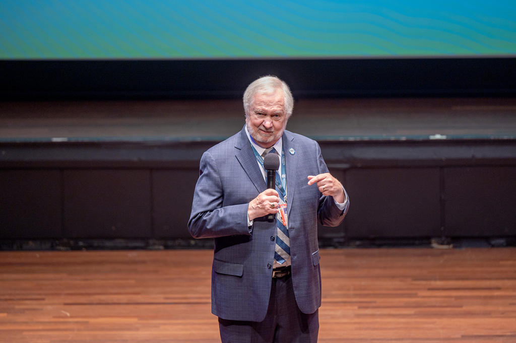 A man in a suit holds a microphone on a stage while speaking to a large crowd.