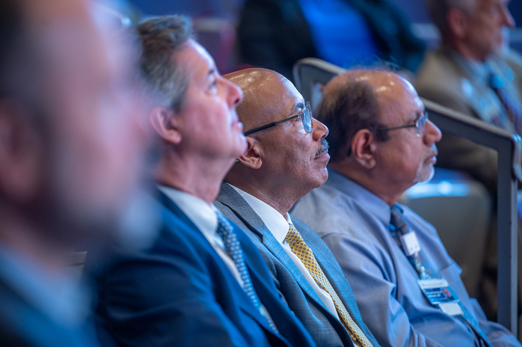 Three men in suits sit as part of a large group listening to a presentation in a large auditorium.