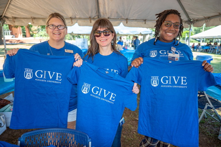 Three women hold up t-shirts with the text IGIVE on them