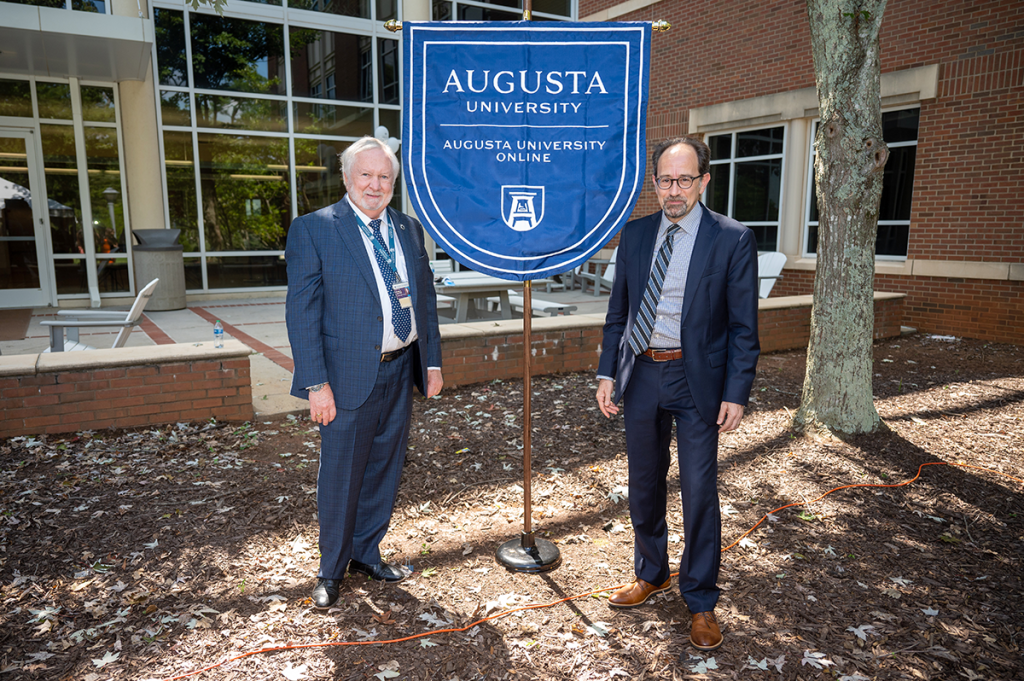 Two men in suits pose for a picture in front of a banner bearing the name and logo of Augusta University Online.