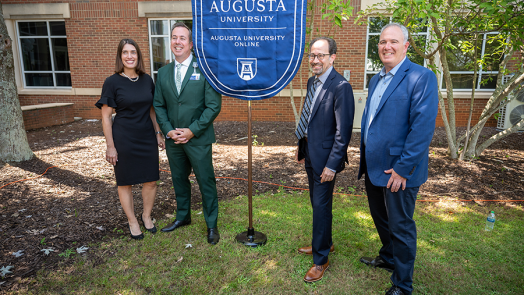 Three men and a woman stand on each side of a flag bearing the logo for Augusta University Online.