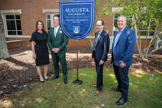Three men and a woman stand on each side of a flag bearing the logo for Augusta University Online.