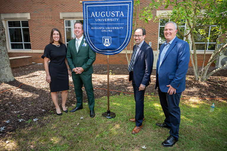 Three men and a woman stand on each side of a flag bearing the logo for Augusta University Online.