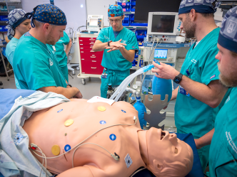 Four medical students in scrubs stand around a simulation dummy while an instructor demonstrates a technique.
