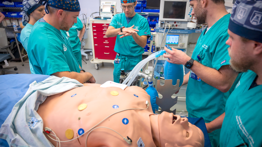 Four medical students in scrubs stand around a simulation dummy while an instructor demonstrates a technique.
