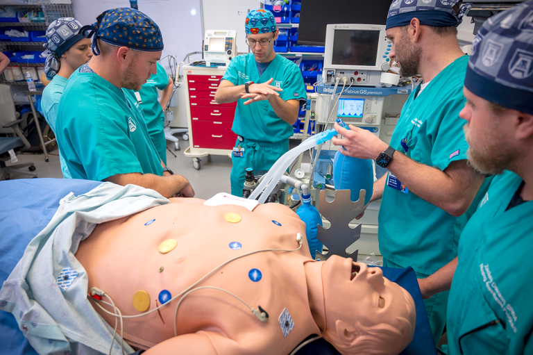 Four medical students in scrubs stand around a simulation dummy while an instructor demonstrates a technique.