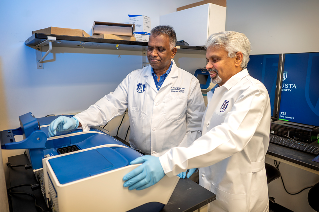 Two men in lab coats stand in front of a piece of lab machinery.