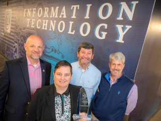 Four people stand in front of a wall with the words "Information Technology" while holding an award.