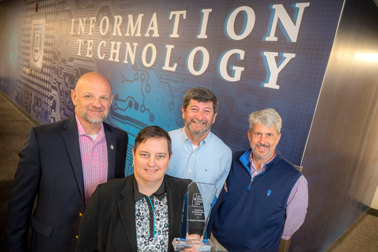 Four people stand in front of a wall with the words "Information Technology" while holding an award.