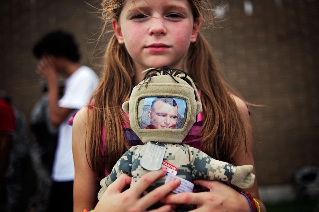 A young girl holds a stuffed doll whith a photo inside a helmet and military dog tags around its neck.