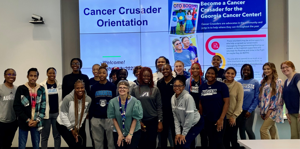 a group of more than 20 men and women pose in front of a screen with the words Cancer Crusader Orientation behind them with information on the screen about the program