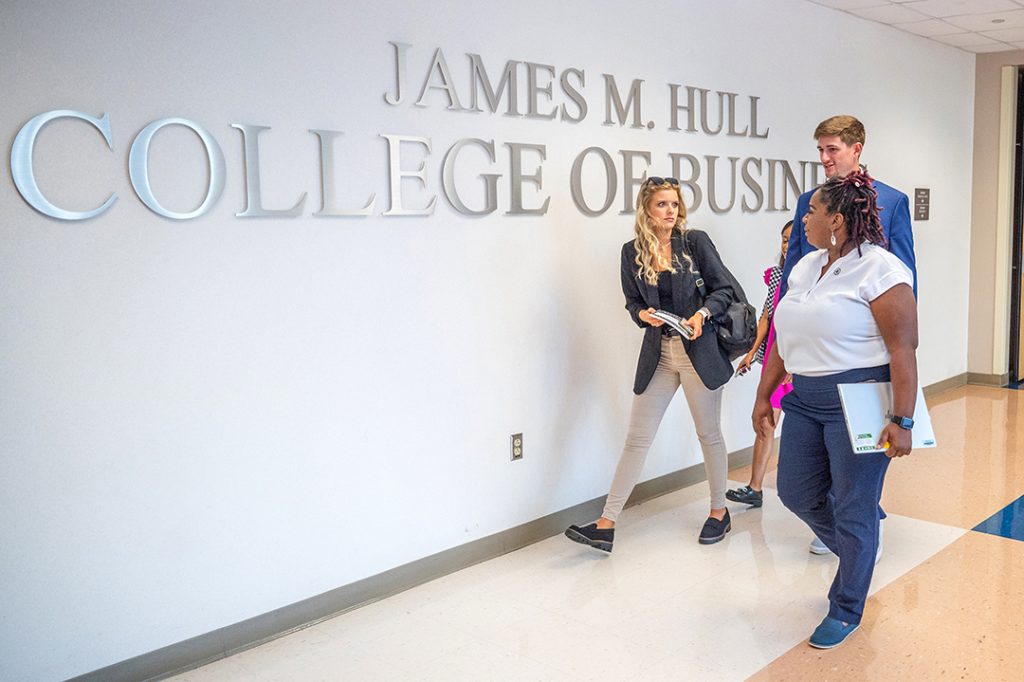 two female and a male student walk past the words James M. Hull College of Business on a wall