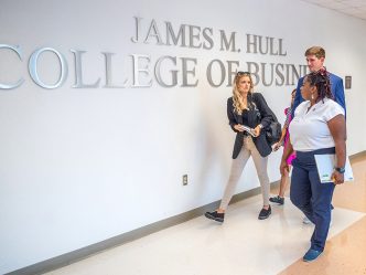 two female and a male student walk past the words James M. Hull College of Business on a wall