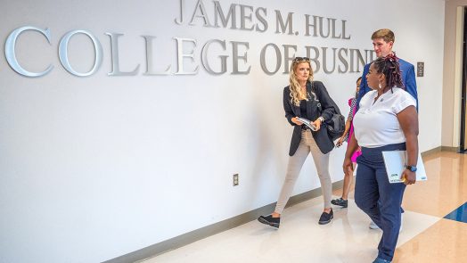 two female and a male student walk past the words James M. Hull College of Business on a wall