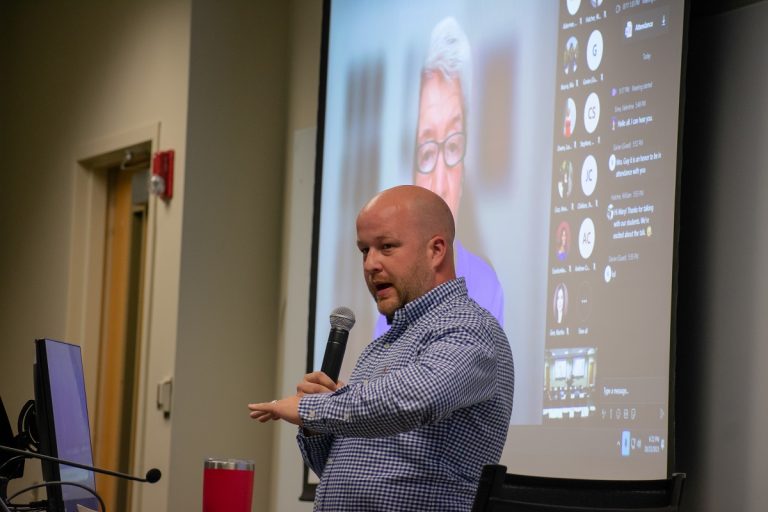 Man talking to a crowd with a zoom interview in the background