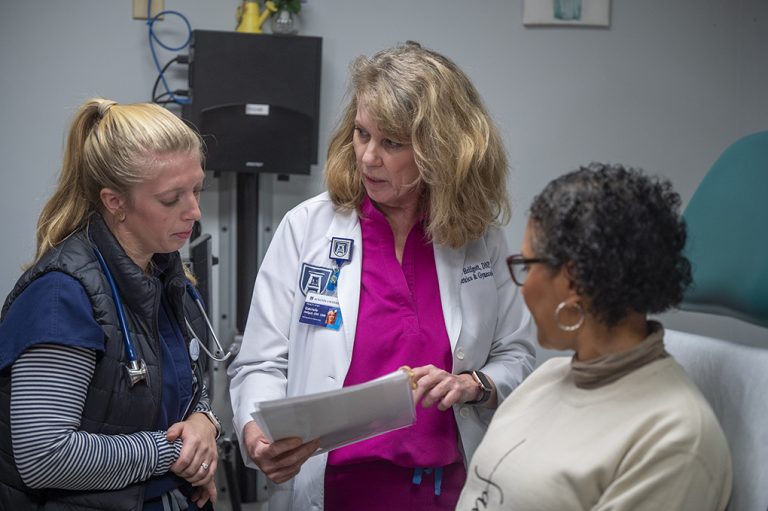 a doctor and a nursing student look at paperwork in an exam room with a woman sitting on the exam table.