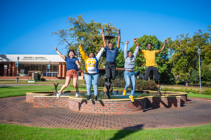five college students jump into the air in front of a fountain