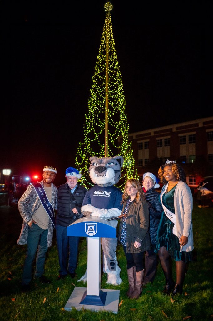 Two men and 3 women surround Augusta University's mascot Augustus behind the switch that lit the Christmas tree they are all standing in front of.