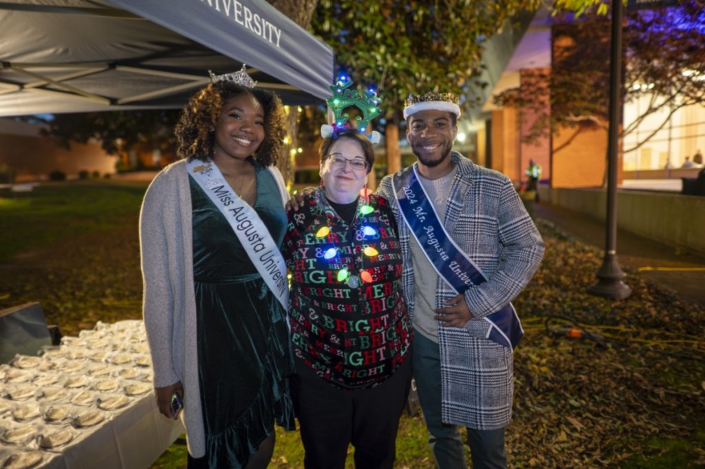 Two women and one man pose for a photo at a Christmas event. The woman on the left is wearing a Miss Augusta University sash, the man on the right is wearing a Mr. Augusta University sash and the woman in the middle is dressed in holiday attire with a lighted handband and necklace.