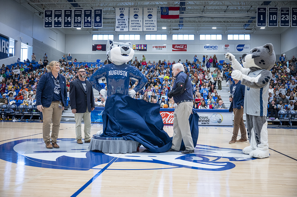 Two men and a woman pull a cover off a statue while an athletics mascot stands nearby.